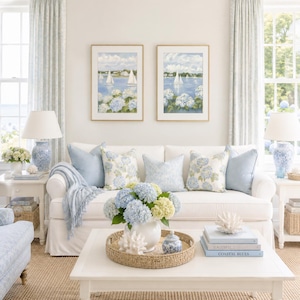 May include: A white sofa with light blue and floral patterned throw pillows, a white coffee table with a round woven tray holding a blue and white hydrangea bouquet, and two framed sailboat paintings hanging on the wall above the sofa.