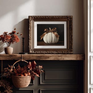 May include: A framed painting of a white pumpkin tied with a brown ribbon, displayed on a wooden mantel. The ornate, dark gold frame surrounds the artwork, which features a dark background. The pumpkin is centered, showcasing its ribbed texture.