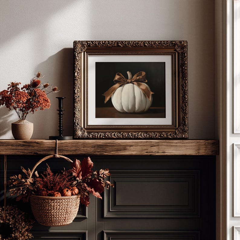 May include: A framed painting of a white pumpkin tied with a brown ribbon, displayed on a wooden mantel. The ornate, dark gold frame surrounds the artwork, which features a dark background. The pumpkin is centered, showcasing its ribbed texture.