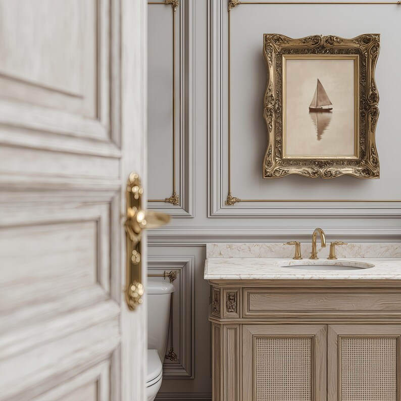 May include: A bathroom scene featuring a framed sailboat painting, a light wood vanity with a marble countertop, and gold fixtures. The walls are gray with decorative molding. A partially open white door with gold hardware is in the foreground.