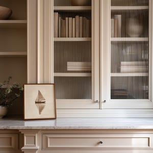 May include: A framed sailboat art print sits on a marble countertop in front of a built-in cabinet with glass doors. The cabinet is filled with books. The color scheme is neutral with beige and white tones.