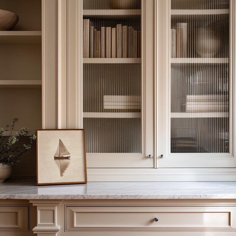 May include: A framed sailboat art print sits on a marble countertop in front of a built-in cabinet with glass doors. The cabinet is filled with books. The color scheme is neutral with beige and white tones.