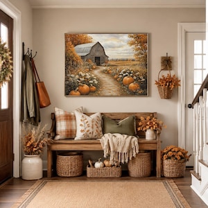 May include: A wooden entryway bench with three throw pillows, a cream-colored throw blanket, and three woven baskets underneath. A framed landscape painting of a barn and pumpkins hangs above the bench, flanked by a coat rack and a wall basket.