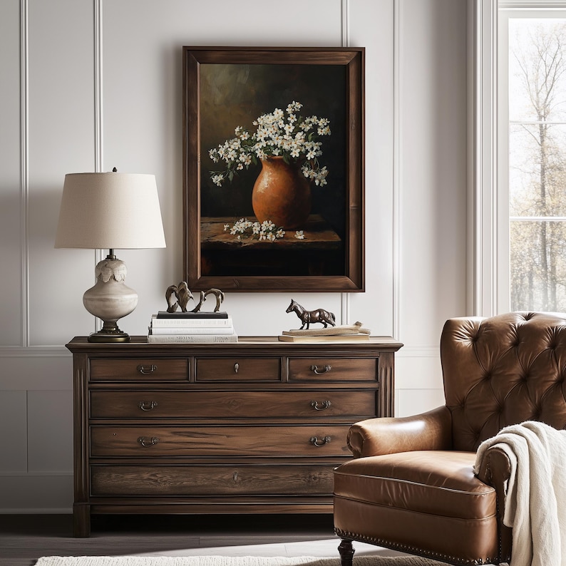 May include: A framed oil painting depicting a terracotta vase filled with white blossoms resting on a wooden surface. The artwork is displayed above a dark wood dresser featuring six drawers, a table lamp with a cream shade, and two small horse figurines.