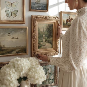 May include: A gallery wall featuring multiple framed artworks, including a central painting of a figure in a garden within an ornate gold frame, a butterfly print, a landscape with ducks, a seascape, and a horse rider, all arranged on a white wall.