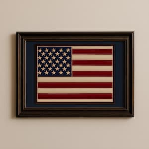 May include: Framed American flag with a dark brown wooden frame. The flag features red and white stripes, a blue field with white stars, and a cream-colored border. The flag is displayed on a navy blue background.