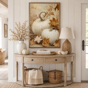 May include: A semi-circular wooden console table holds a white vase with dried branches, a small white pumpkin, a woven tray with books, a candle, and a woven lamp. A framed print of white pumpkins and autumn leaves hangs above the table.