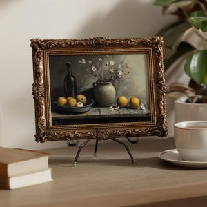 May include: A rectangular painting in an ornate, dark gold-toned frame resting on a metal easel. The artwork depicts a still life scene featuring a dark bottle, a ceramic vase with white flowers, and several yellow fruits on a wooden surface.