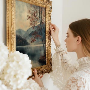 May include: A rectangular landscape painting in an ornate, heavy gold-toned frame. The artwork depicts a serene lake surrounded by misty mountains and a single tree with reddish-brown foliage. The frame features intricate, raised floral carvings.