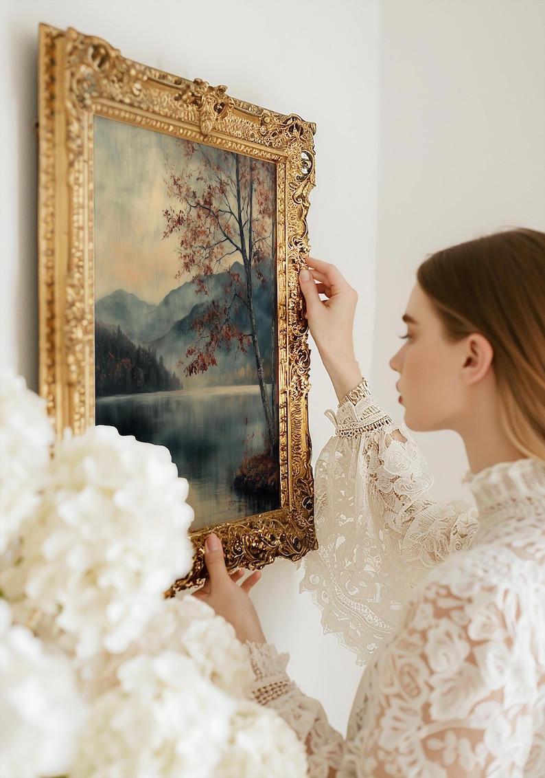 May include: A rectangular landscape painting in an ornate, heavy gold-toned frame. The artwork depicts a serene lake surrounded by misty mountains and a single tree with reddish-brown foliage. The frame features intricate, raised floral carvings.
