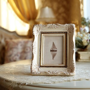 May include: An ornate, off-white picture frame with a gold inner border. The frame holds a sepia-toned image of a sailboat on calm water. The frame sits on a lace-covered table, with a blurred background.