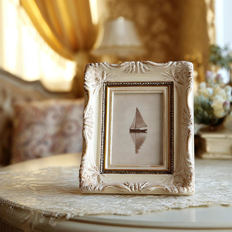 May include: An ornate, off-white picture frame with a gold inner border. The frame holds a sepia-toned image of a sailboat on calm water. The frame sits on a lace-covered table, with a blurred background.