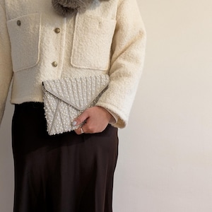 May include: Cream-colored jacket with gold buttons and two front pockets. A silver beaded clutch is held in front of a dark brown skirt. A gray faux fur scarf is worn around the neck. The outfit is neutral in color.