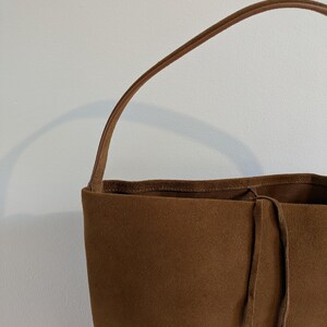 K&ouml;nnte beinhalten: Eine braune Wildleder-Tragetasche mit einem einzelnen Henkel. Die Tasche hat einen Kordelzugverschluss und ein weiches, strukturiertes Aussehen. Der Griff ist in einem helleren Braunton gehalten.