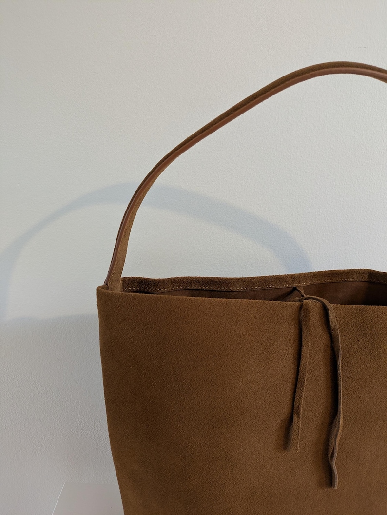 K&ouml;nnte beinhalten: Eine braune Wildleder-Tragetasche mit einem einzelnen Henkel. Die Tasche hat einen Kordelzugverschluss und ein weiches, strukturiertes Aussehen. Der Griff ist in einem helleren Braunton gehalten.