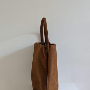 K&ouml;nnte beinhalten: Eine braune Wildleder-Tragetasche mit einem einzelnen Griff. Die Tasche hat eine rechteckige Form mit einem leicht verj&uuml;ngten Design. Der Griff ist oben befestigt und hat die gleiche Farbe wie die Tasche. Die Tasche steht auf einer wei&szlig;en Oberfl&auml;che.