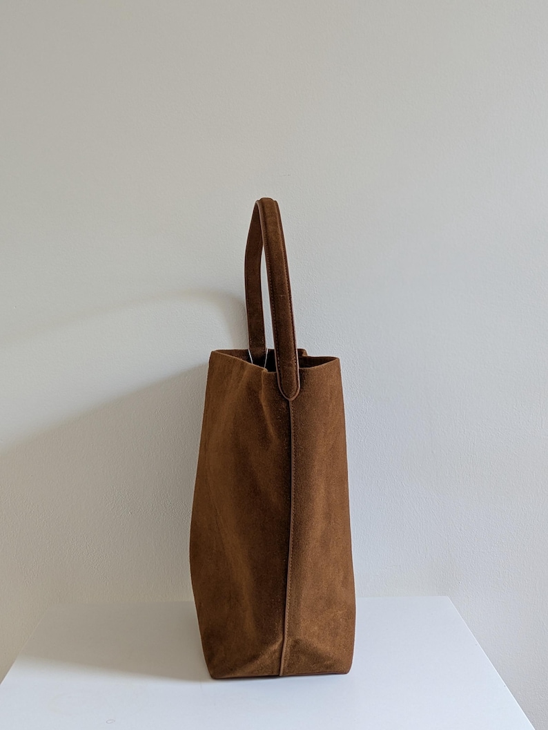 K&ouml;nnte beinhalten: Eine braune Wildleder-Tragetasche mit einem einzelnen Griff. Die Tasche hat eine rechteckige Form mit einem leicht verj&uuml;ngten Design. Der Griff ist oben befestigt und hat die gleiche Farbe wie die Tasche. Die Tasche steht auf einer wei&szlig;en Oberfl&auml;che.