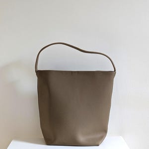 May include: A taupe-coloured bucket bag with a textured surface and a single arched shoulder strap. The bag features a wide, open top and a structured, tapered base, presenting a clean, simple silhouette without any visible hardware or external pockets.