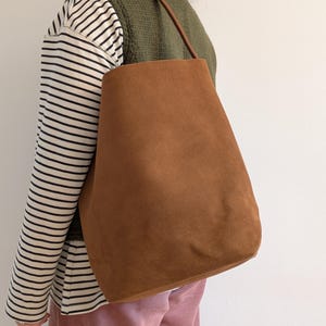 K&ouml;nnte beinhalten: Eine braune Wildleder-Tragetasche mit einem einzelnen Schulterriemen. Die Tasche hat eine warme, erdige braune Farbe und ein schlichtes, minimalistisches Design. Der Riemen ist dunkler braun. Die Tasche wird &uuml;ber der Schulter getragen.