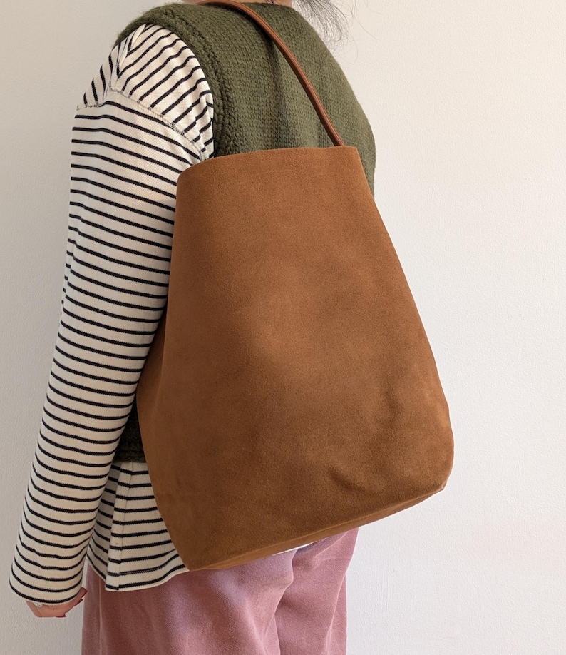 K&ouml;nnte beinhalten: Eine braune Wildleder-Tragetasche mit einem einzelnen Schulterriemen. Die Tasche hat eine warme, erdige braune Farbe und ein schlichtes, minimalistisches Design. Der Riemen ist dunkler braun. Die Tasche wird &uuml;ber der Schulter getragen.