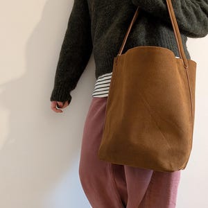 K&ouml;nnte beinhalten: Eine braune Wildleder-Tragetasche mit langen, d&uuml;nnen Tr&auml;gern. Die Tasche ist rechteckig mit einer leicht gebogenen Oberkante. Die Tr&auml;ger sind hellbraun. Die Tasche wird von einer Person getragen, die einen dunkelgr&uuml;nen Pullover und eine rosa Hose tr&auml;gt.