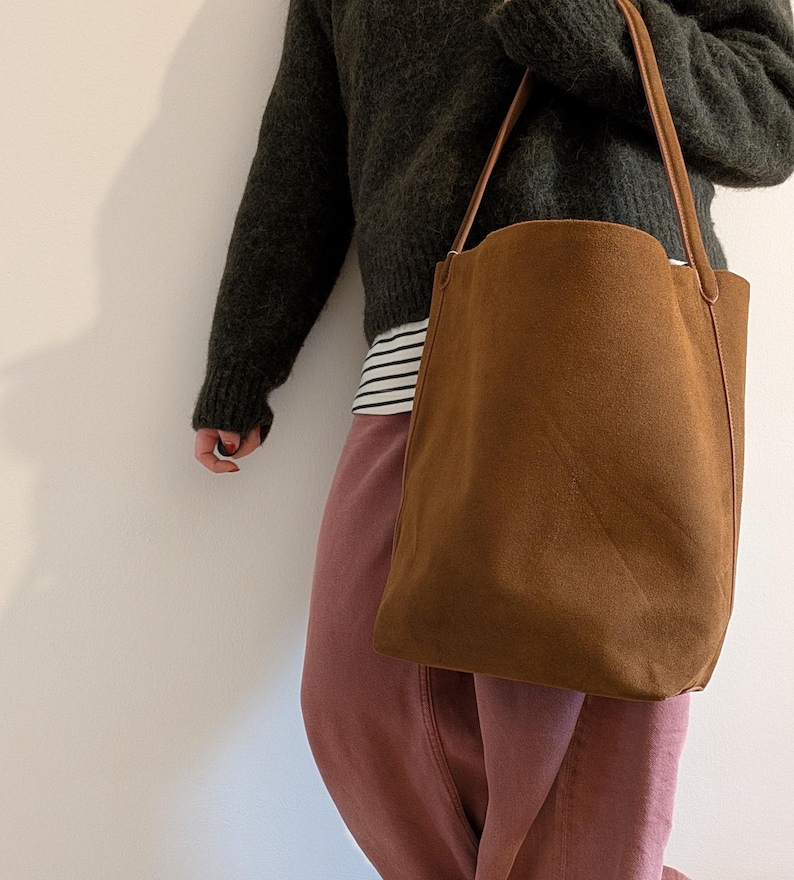 K&ouml;nnte beinhalten: Eine braune Wildleder-Tragetasche mit langen, d&uuml;nnen Tr&auml;gern. Die Tasche ist rechteckig mit einer leicht gebogenen Oberkante. Die Tr&auml;ger sind hellbraun. Die Tasche wird von einer Person getragen, die einen dunkelgr&uuml;nen Pullover und eine rosa Hose tr&auml;gt.