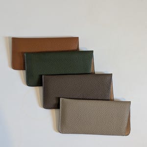 May include: Four leather eyeglass cases in shades of brown, green, and grey are arranged diagonally. Each case has a textured surface and a rectangular shape with a flap closure. The cases are displayed on a white background.