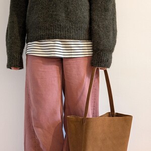K&ouml;nnte beinhalten: Eine Person tr&auml;gt einen dunkelgr&uuml;nen Pullover &uuml;ber einem gestreiften Hemd und einer rosa Hose und h&auml;lt eine braune Tragetasche. Die Tasche hat lange Griffe und eine rechteckige Form. Das Outfit ist l&auml;ssig und stilvoll.