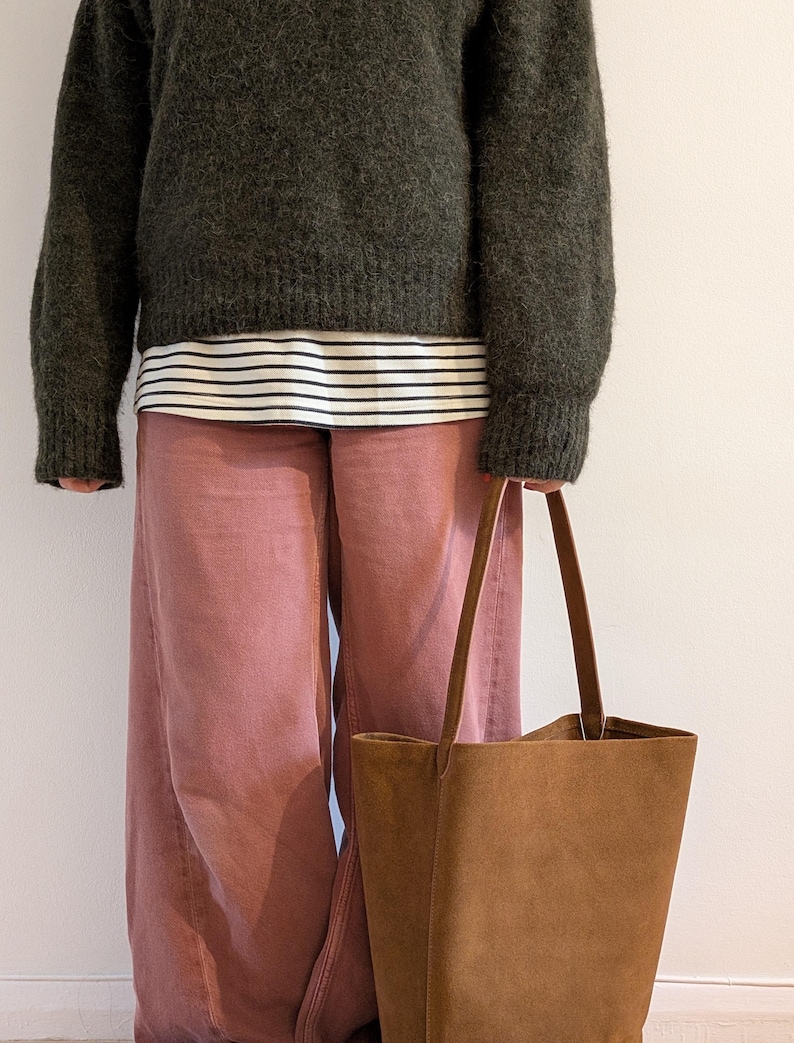 K&ouml;nnte beinhalten: Eine Person tr&auml;gt einen dunkelgr&uuml;nen Pullover &uuml;ber einem gestreiften Hemd und einer rosa Hose und h&auml;lt eine braune Tragetasche. Die Tasche hat lange Griffe und eine rechteckige Form. Das Outfit ist l&auml;ssig und stilvoll.