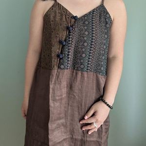 Peut inclure: Robe sans manches marron et à motifs avec fines bretelles. La partie supérieure présente un imprimé floral et géométrique, fermé par des boutons bleu foncé. La moitié inférieure est de couleur marron uni.