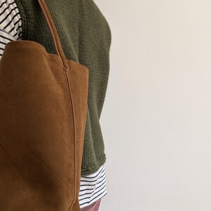 K&ouml;nnte beinhalten: Eine braune Wildleder-Tragetasche mit langem Henkel. Die Tasche wird &uuml;ber der Schulter getragen. Die Person tr&auml;gt einen gr&uuml;nen Pullover &uuml;ber einem gestreiften Hemd und einer rosa Hose. Der Hintergrund ist eine schlichte wei&szlig;e Wand.