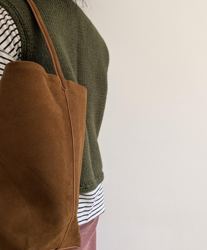 K&ouml;nnte beinhalten: Eine braune Wildleder-Tragetasche mit langem Henkel. Die Tasche wird &uuml;ber der Schulter getragen. Die Person tr&auml;gt einen gr&uuml;nen Pullover &uuml;ber einem gestreiften Hemd und einer rosa Hose. Der Hintergrund ist eine schlichte wei&szlig;e Wand.