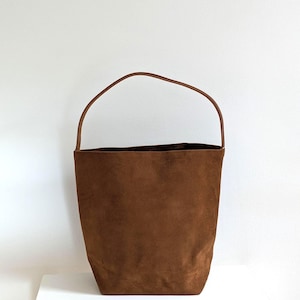 K&ouml;nnte beinhalten: Eine braune Wildleder-Eimertasche mit einem einzelnen Griff. Die Tasche hat eine zylindrische Form und eine glatte Textur. Der Griff hat einen dunkleren Braunton. Die Tasche steht auf einer wei&szlig;en Oberfl&auml;che vor einem wei&szlig;en Hintergrund.