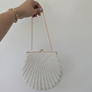 May include: A white beaded handbag shaped like a seashell with a gold-toned metal frame, a kiss-lock clasp, and a thin gold-toned chain strap. The exterior is covered in small white beads arranged in a radiating fan pattern across the surface.