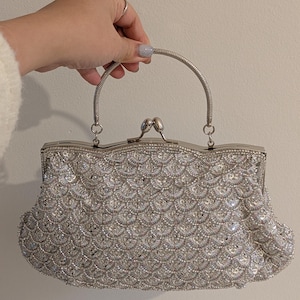 Puede incluir: Un bolso de mano plateado con un patrón de escamas cubierto de cuentas y lentejuelas iridiscentes. El bolso tiene un marco metálico, un cierre de boquilla y una sola asa plateada redondeada sujeta a la parte superior con anillas.