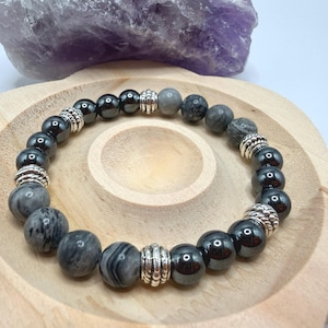 May include: A bracelet with dark grey and patterned grey beads, and silver-coloured metal spacers. The bracelet is presented on a wooden tray, with a large purple crystal in the background.