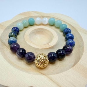 May include: A beaded bracelet featuring a variety of colorful gemstones, including blue, purple, and white beads. A gold-colored filigree bead is centered in the bracelet. The bracelet is displayed on a light wooden plate.