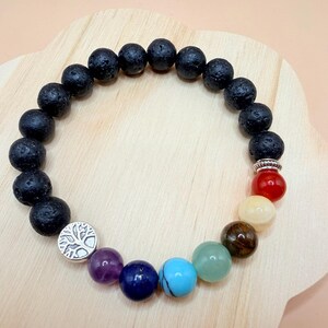 May include: A bracelet with black lava rock beads and chakra-coloured gemstones. The bracelet includes a silver charm with a tree of life design. The gemstones are in shades of purple, blue, turquoise, green, yellow, and red.