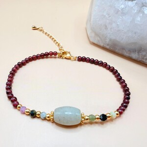 May include: A bracelet with a central light blue, oval-shaped stone, small, round, dark red beads, and gold-coloured accents. The bracelet includes colourful beads. The clasp is gold-coloured. The bracelet is likely to be approximately 17-20 cm in length.