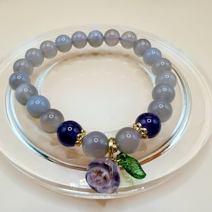 May include: A bracelet with grey and navy blue beads. The bracelet has a charm with a purple flower and a green leaf. The bracelet is displayed on a clear glass plate.