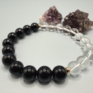 May include: A bracelet with alternating black and clear quartz beads. The beads are strung on a clear elastic cord, with gold-coloured spacer beads. Two rough-cut gemstones are in the background.