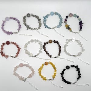 May include: An assortment of gemstone bracelets in various colors, including purple, gray, blue, and yellow. Each bracelet features small, irregularly shaped stones strung together with a silver-colored chain and clasp. The bracelets are displayed on a white surface.