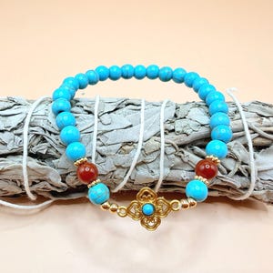 Peut inclure: Un bracelet de perles turquoise avec des détails rouges et dorés. Le bracelet est orné d'un fermoir en filigrane doré avec une pierre turquoise. Le bracelet est présenté sur un fagot de sauge blanche.