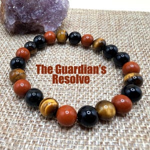 Peut inclure: Un bracelet de perles avec un mélange de pierres précieuses rondes et polies, de couleurs noir, brun-rouge et œil de tigre. Le texte "The Guardian's Resolve" est superposé à l'image. Un géode d'améthyste est en arrière-plan.