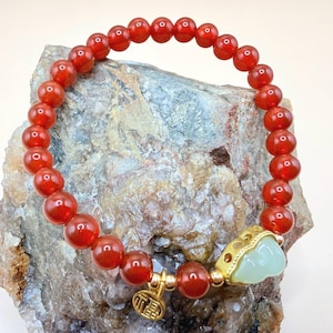 May include: A red beaded bracelet with a gold charm and a light green jade accent. The bracelet is made of round, polished red beads and is displayed on a textured rock surface. A small gold charm with Chinese characters hangs from the bracelet.