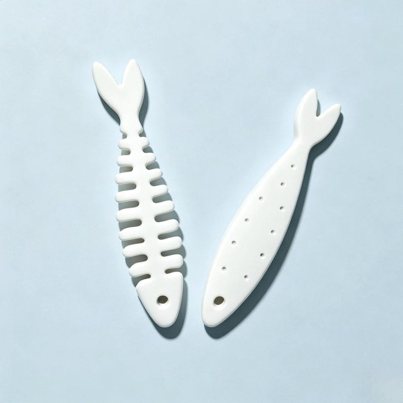 Puede incluir: Dos objetos de cer&aacute;mica blanca con forma de pez. Uno presenta un dise&ntilde;o de espina de pescado con una cola bifurcada y un orificio circular en la cabeza. El segundo objeto es un pez s&oacute;lido con una serie de peque&ntilde;as hendiduras circulares.