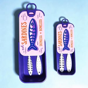 Puede incluir: Dos recipientes rectangulares de color morado con tapas deslizantes que presentan un panel superior rosa claro. Las tapas muestran un gr&aacute;fico de pez azul oscuro, el texto "not SARDINES" y "SKINLESS + BONELESS" y una leng&uuml;eta de apertura.