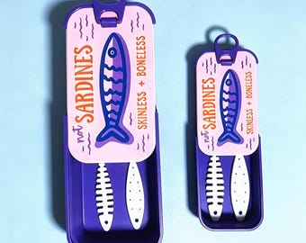 Puede incluir: Dos recipientes rectangulares de color morado con tapas deslizantes que presentan un panel superior rosa claro. Las tapas muestran un gráfico de pez azul oscuro, el texto "not SARDINES" y "SKINLESS + BONELESS" y una lengüeta de apertura.