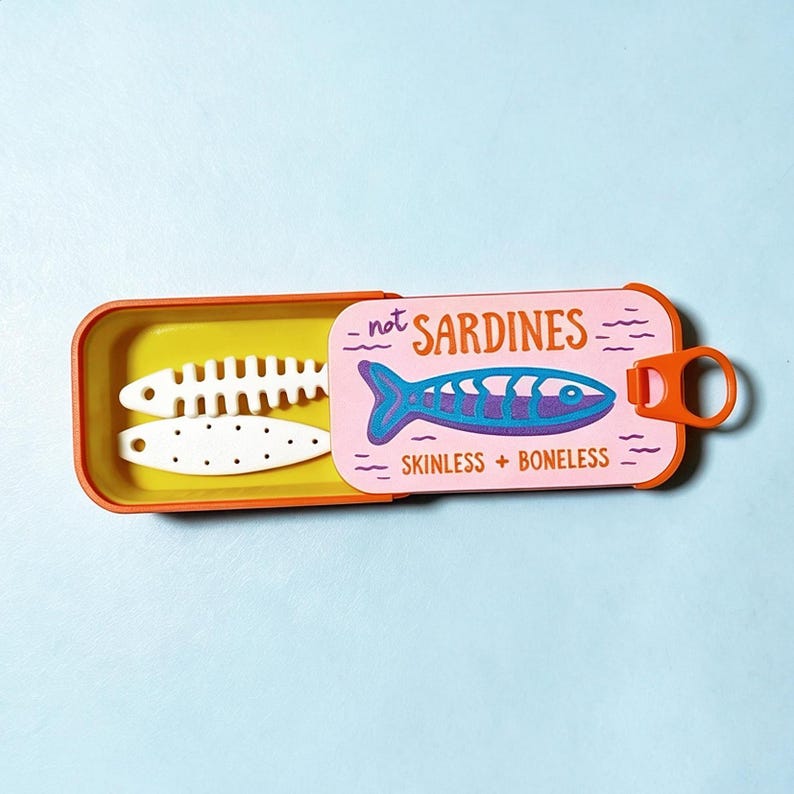 Puede incluir: Una lata rectangular naranja con tapa deslizante que presenta un fondo rosa, una ilustraci&oacute;n de un pez azul y el texto "not SARDINES SKINLESS + BONELESS". Dentro de la lata, forrada en amarillo, hay dos objetos blancos planos con forma de pez.