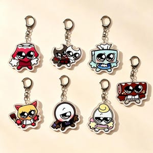 May include: A collection of seven acrylic keychains featuring cartoon characters with large eyes. The designs include a red lamp, two small figures, a blue tissue box, a yellow cat-like creature, a round white character, a pale figure, and a red roll.